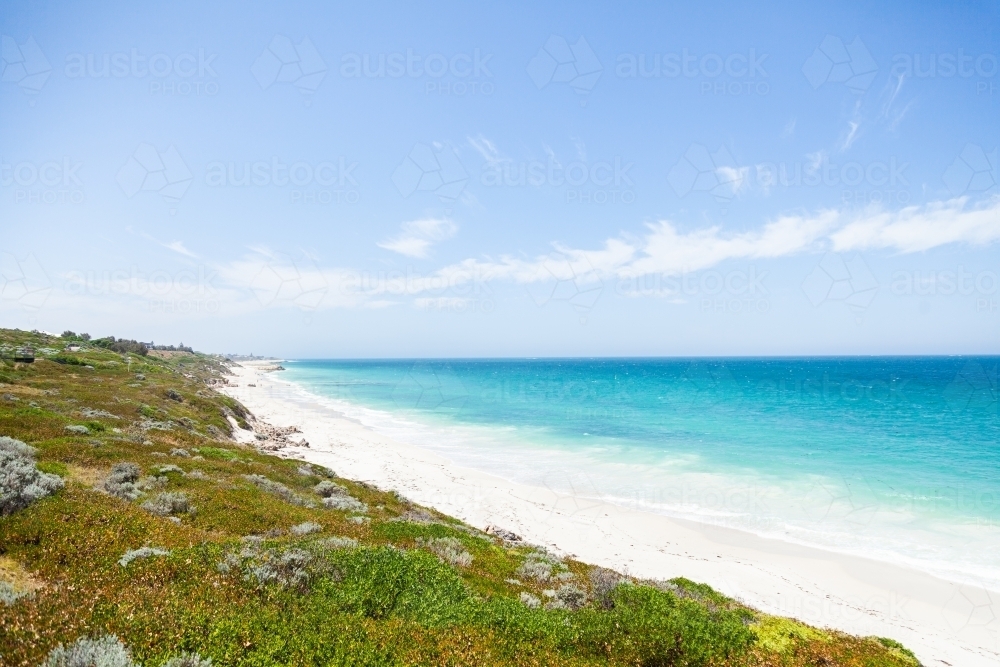 Sunlit coastland with view of shrubs, beach and clear blue coastal waters - Australian Stock Image
