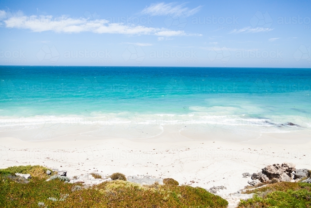 Sunlit coastland with view of shrubs, beach and clear blue coastal waters - Australian Stock Image