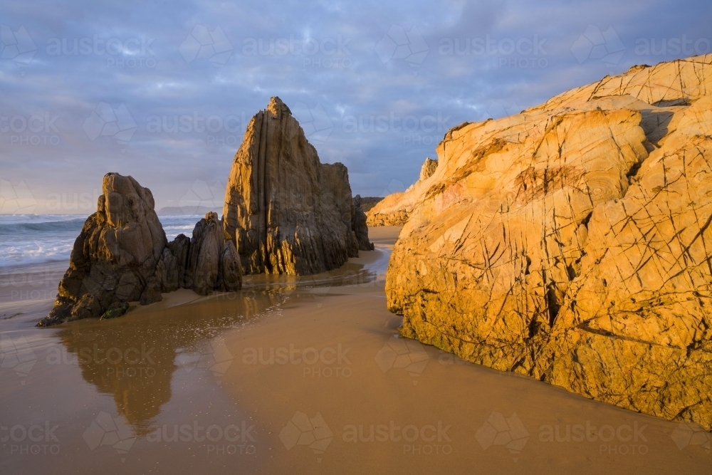 Sunlit yellow coastal rocks at sunrise with sand and cloud pattern - Australian Stock Image