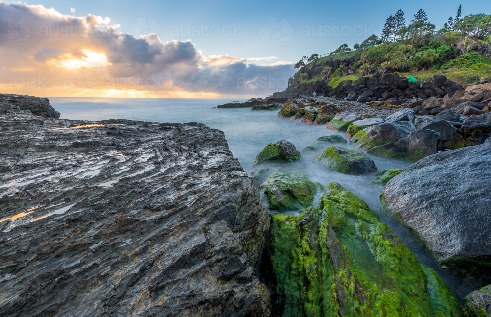 Sunrise at beach with waves crashing over green mossy rocks - Australian Stock Image