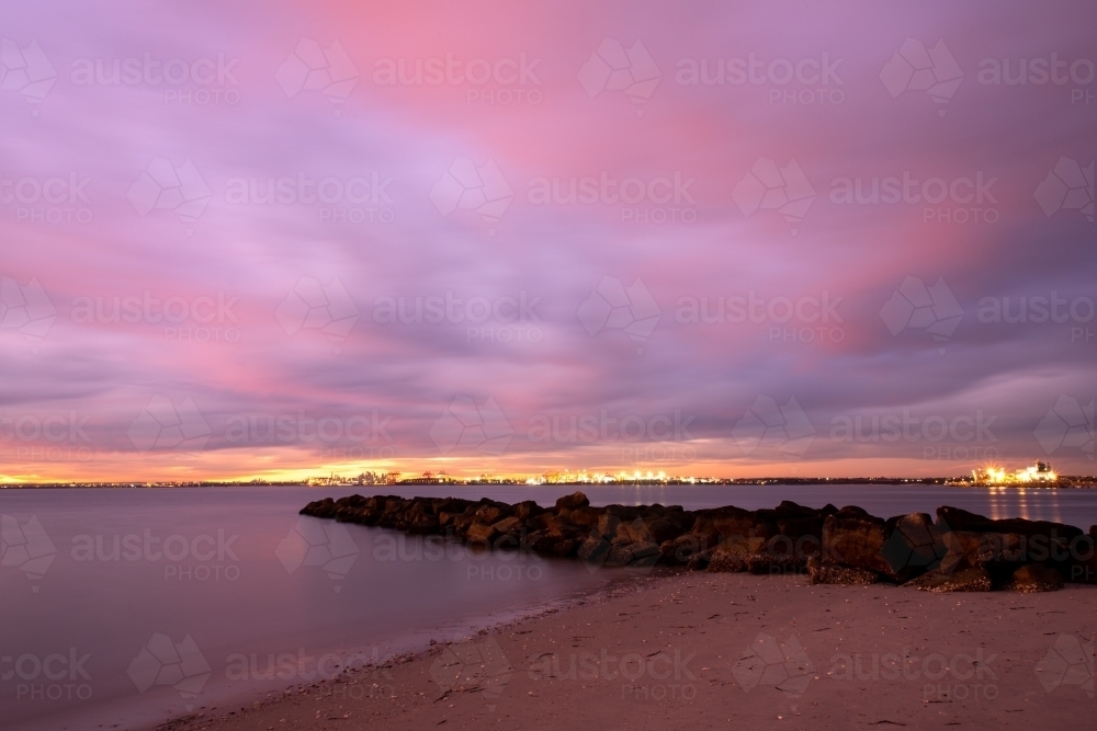 Sunset with lights from Port Botany in the distance and a beach with rock groyne in the foreground - Australian Stock Image