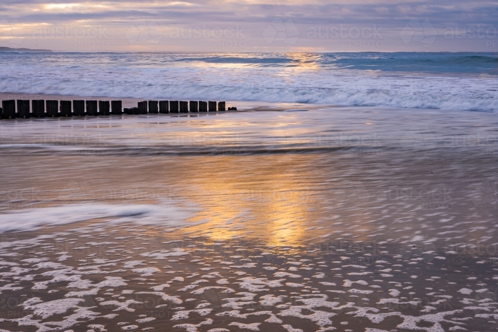 Sunshine reflecting on a wet sandy beach with a wooden groyne and waves in the background - Australian Stock Image