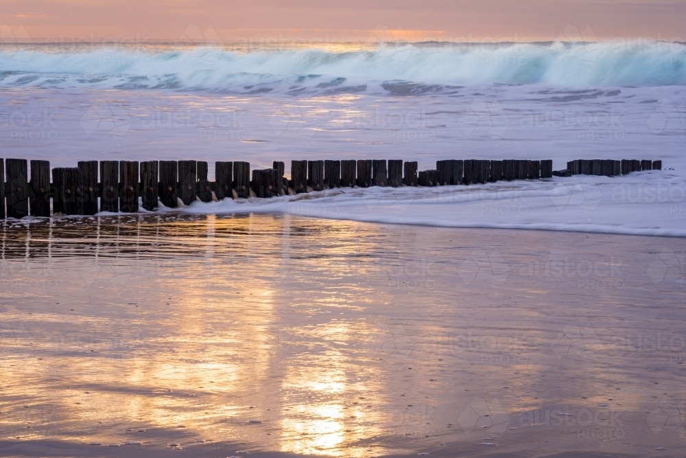 Sunshine reflecting on a wet sandy beach with a wooden groyne and waves in the background - Australian Stock Image