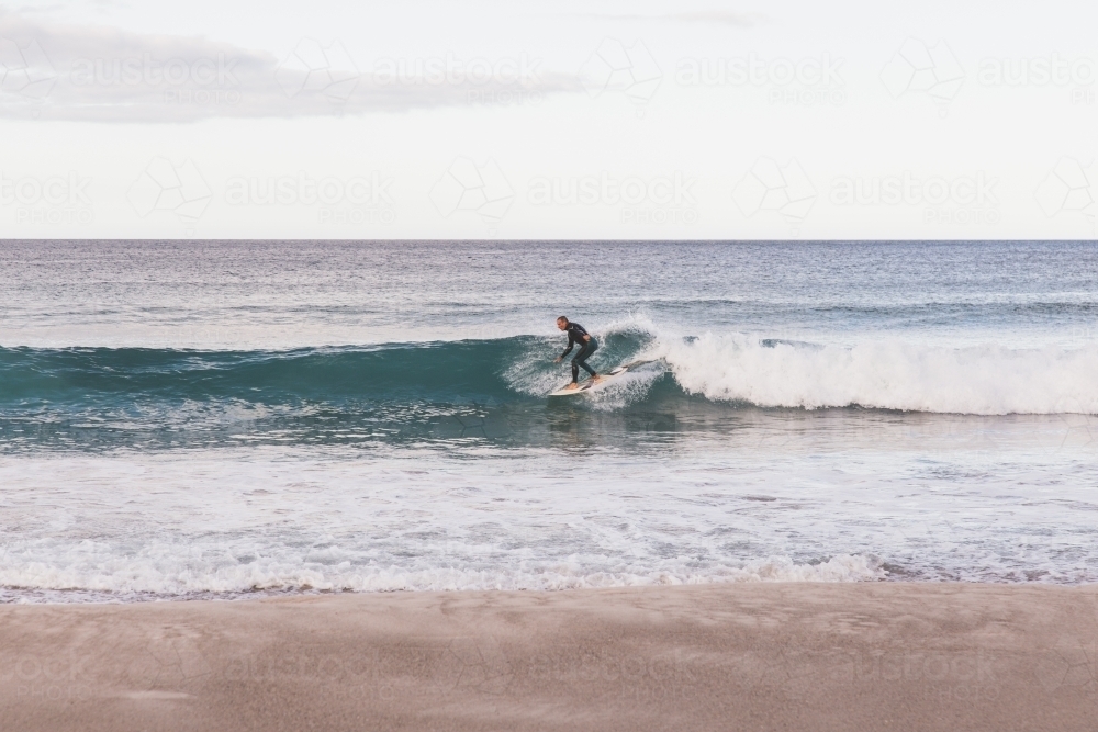 Surfer catching a wave in the ocean, light blue sky and grey cloud and sand in the foreground - Australian Stock Image