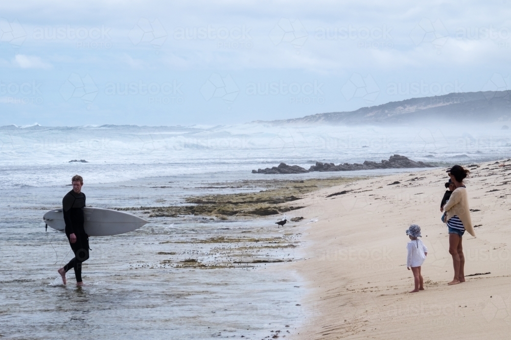 Surfer exiting ocean with young family waiting on the rugged remote beach shoreline - Australian Stock Image