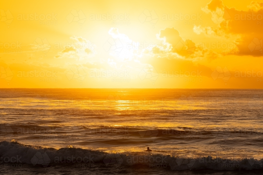 Image of Surfer paddling out into vibrant yellow sunrise over the ocean ...