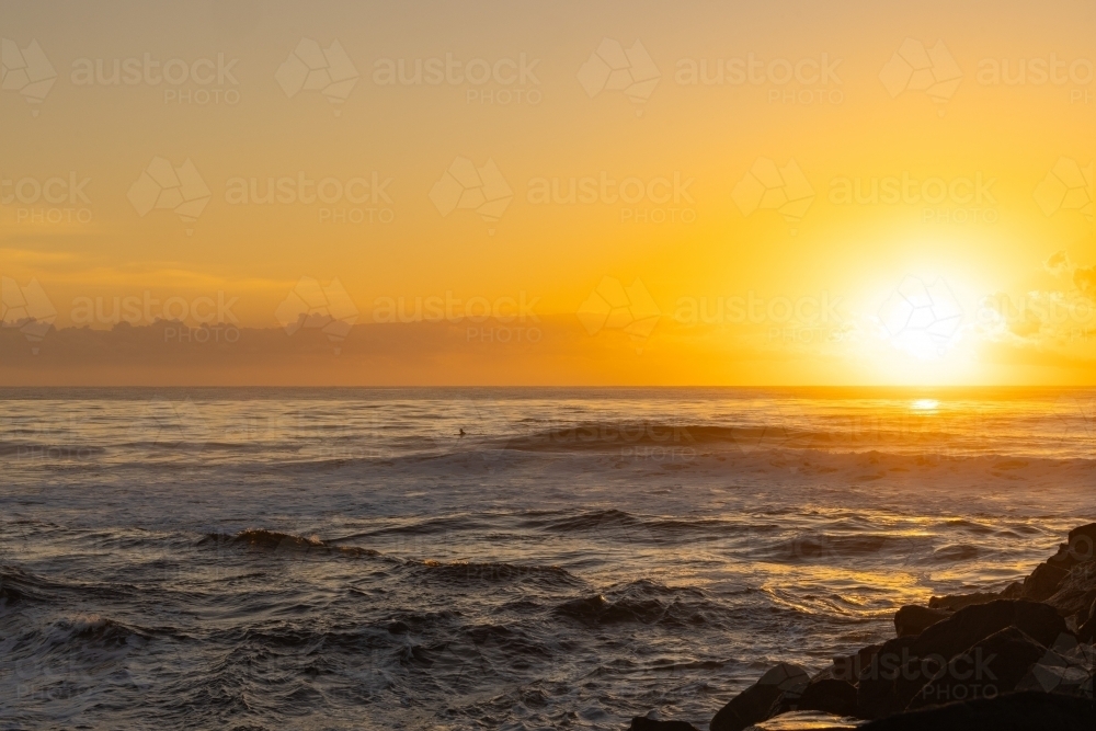 Surfer paddling out into vibrant yellow sunrise over the ocean at Brunswick Heads, NSW - Australian Stock Image