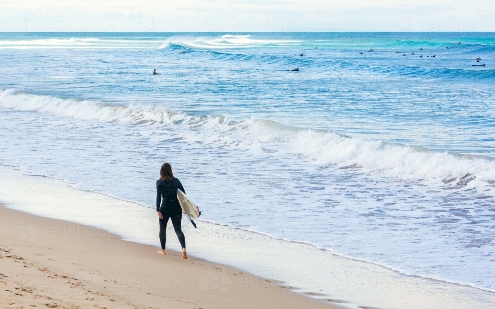Surfer walking along beach with distant people surfing - Australian Stock Image