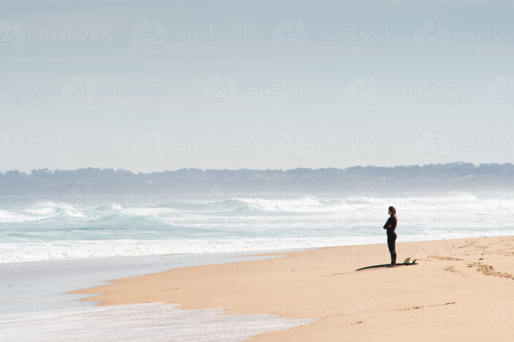 Surfer with board standing on beach looking out to the ocean - Australian Stock Image