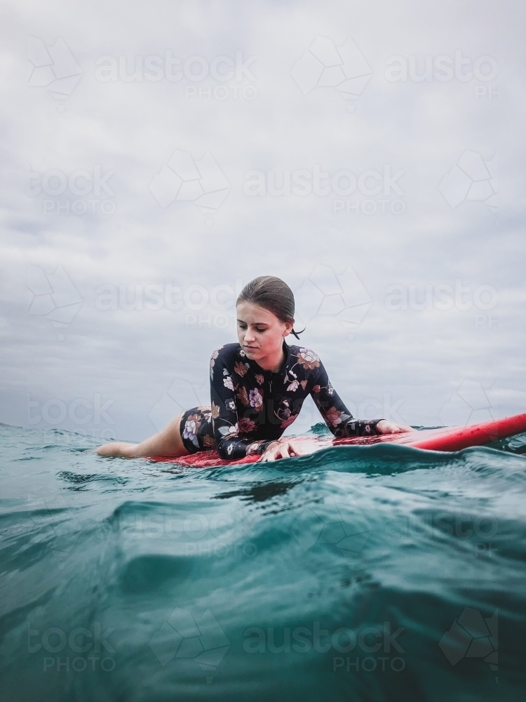 Surfing Teenage girl lying on red surfboard in ocean wearing floral swimsuit on overcast day - Australian Stock Image