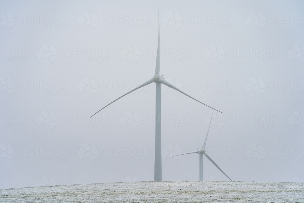 Tall wind turbines in a foggy sky on a snow covered hilltop - Australian Stock Image