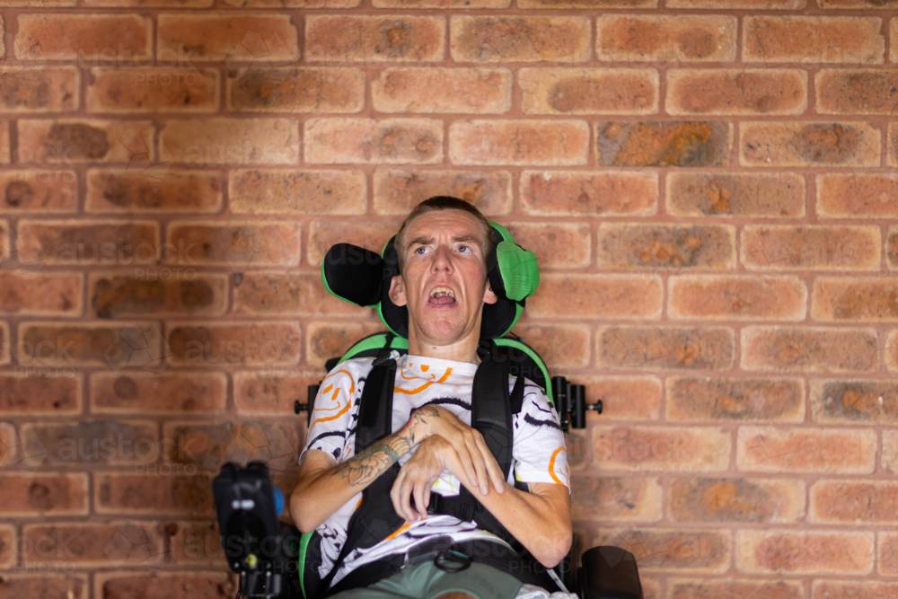 tattooed man sitting in electric wheelchair alone in front of a brick wall - Australian Stock Image
