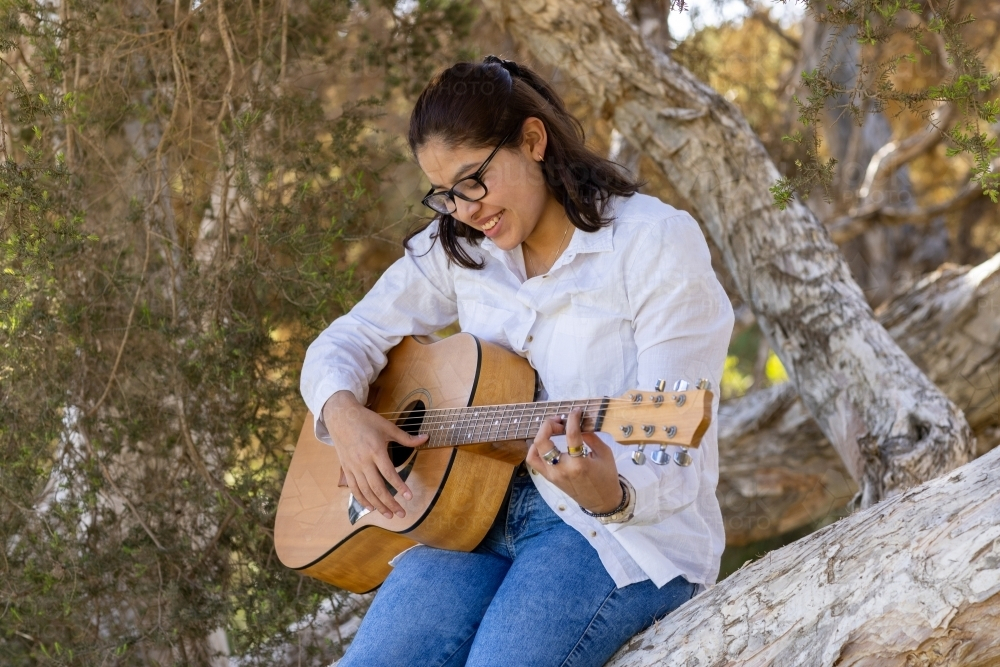 teeen girl sitting in shade of trees strumming a tune on an acoustic guitar - Australian Stock Image