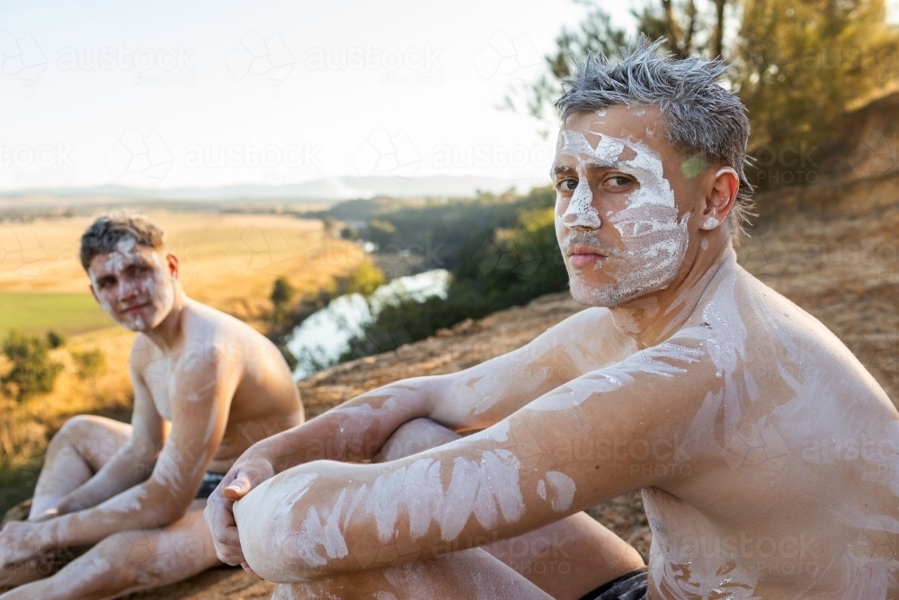 Teen aboriginal cousins in ochre body paint sitting on cliff overlooking australian landscape - Australian Stock Image