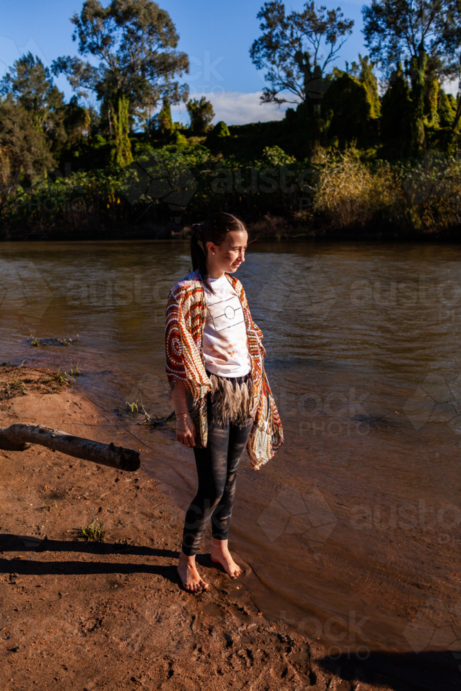 Teen aboriginal girl standing in sunlight by water of inland Australian river - Australian Stock Image