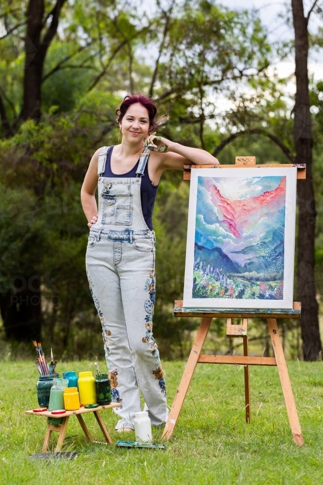 Teen artist leaning on completed painting on easel outside - Australian Stock Image