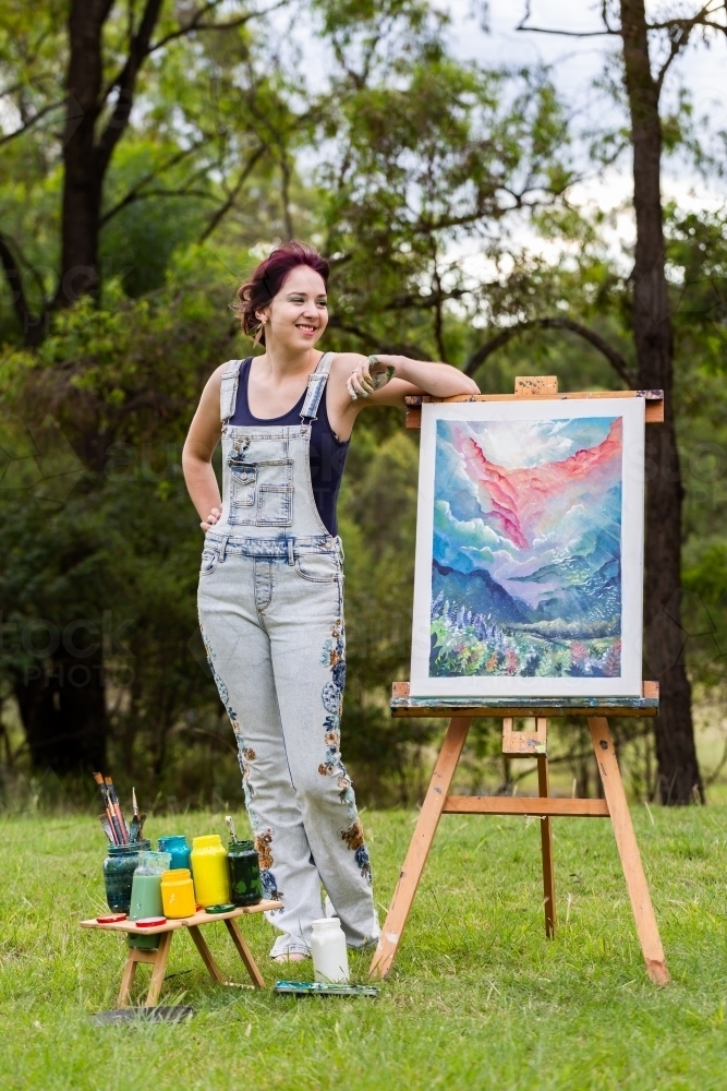 Teen artist leaning on completed painting on easel outside - Australian Stock Image