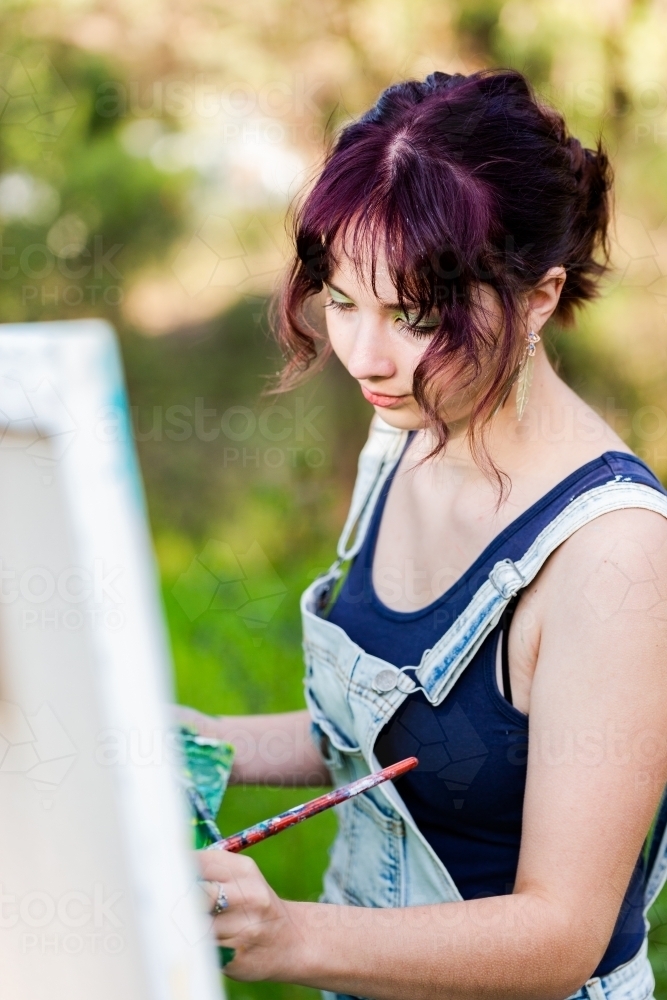 Teen artist with purple hair working on painting in overalls - Australian Stock Image