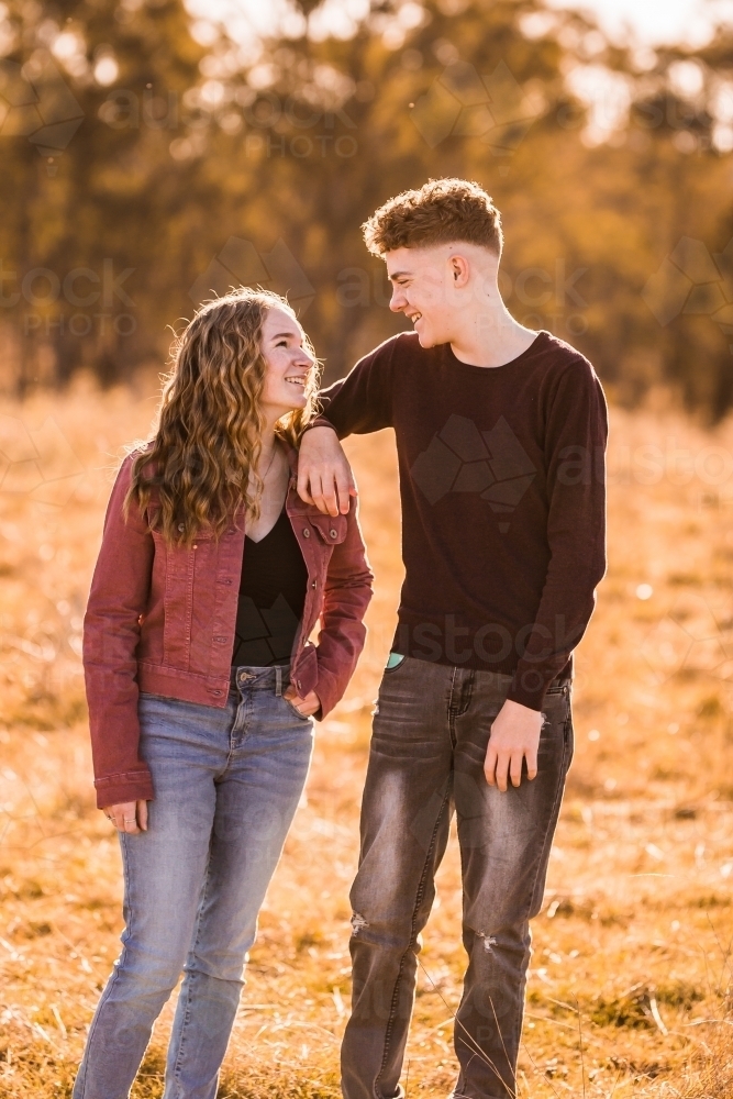 Teen boy and girl looking at each while boy leans on girl other smiling outside - Australian Stock Image