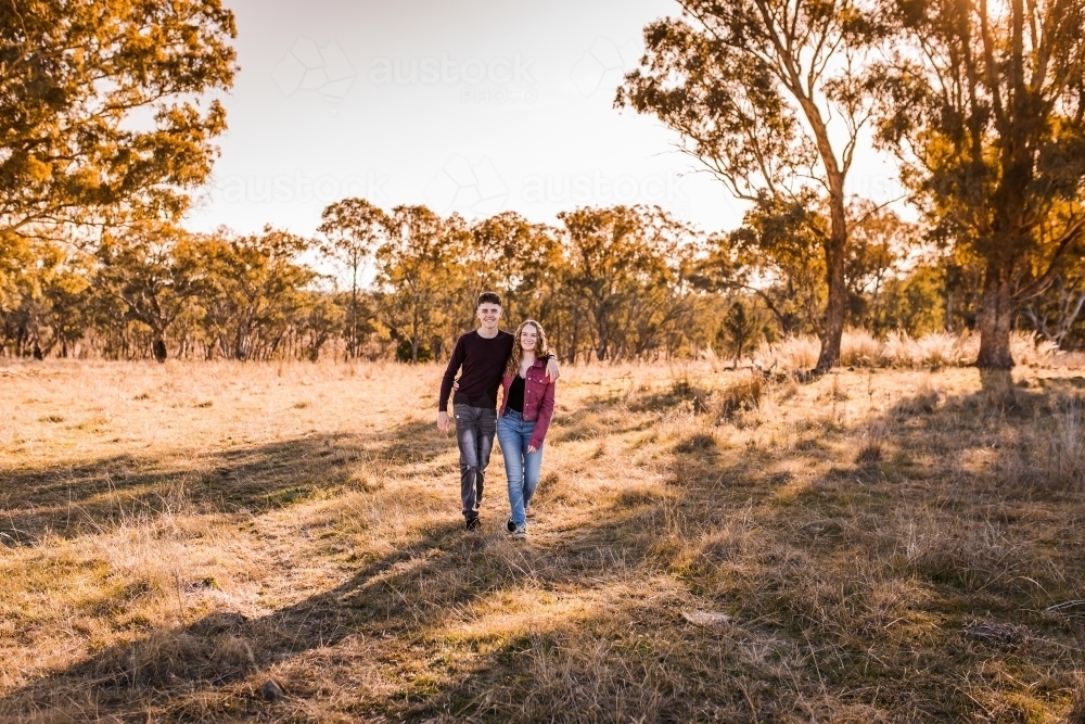 Teen boy and girl walking through paddock arm in arm smiling happy - Australian Stock Image