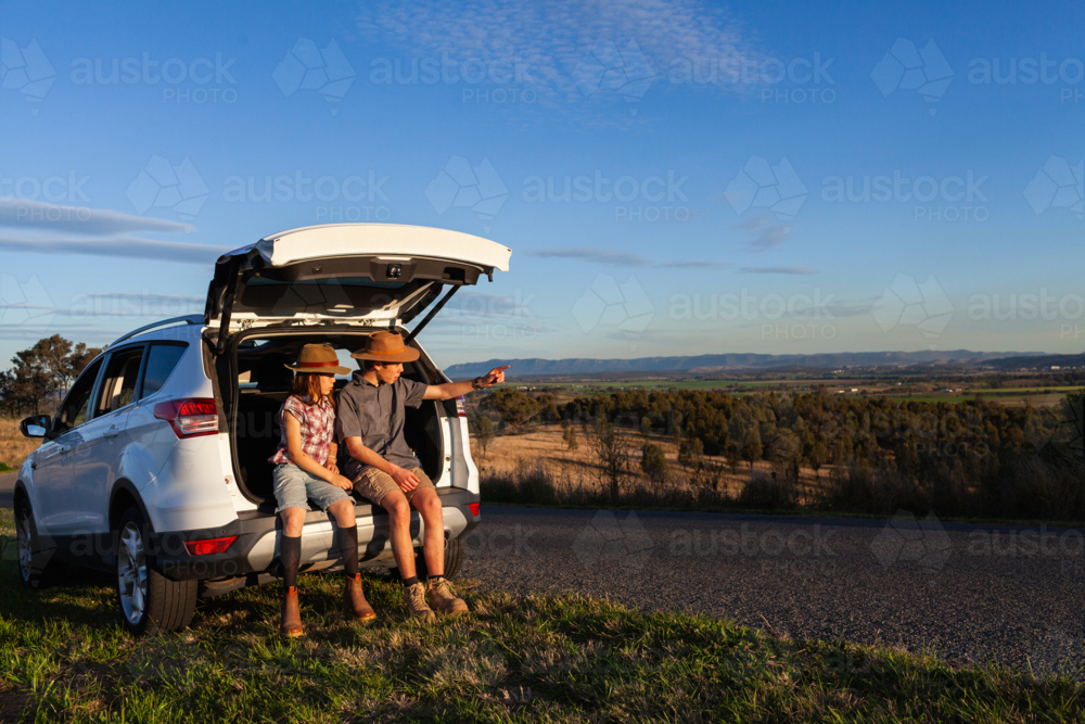 Teen boy and preteen girl siblings sitting in car boot beside rural Australian road in warm sunlight - Australian Stock Image