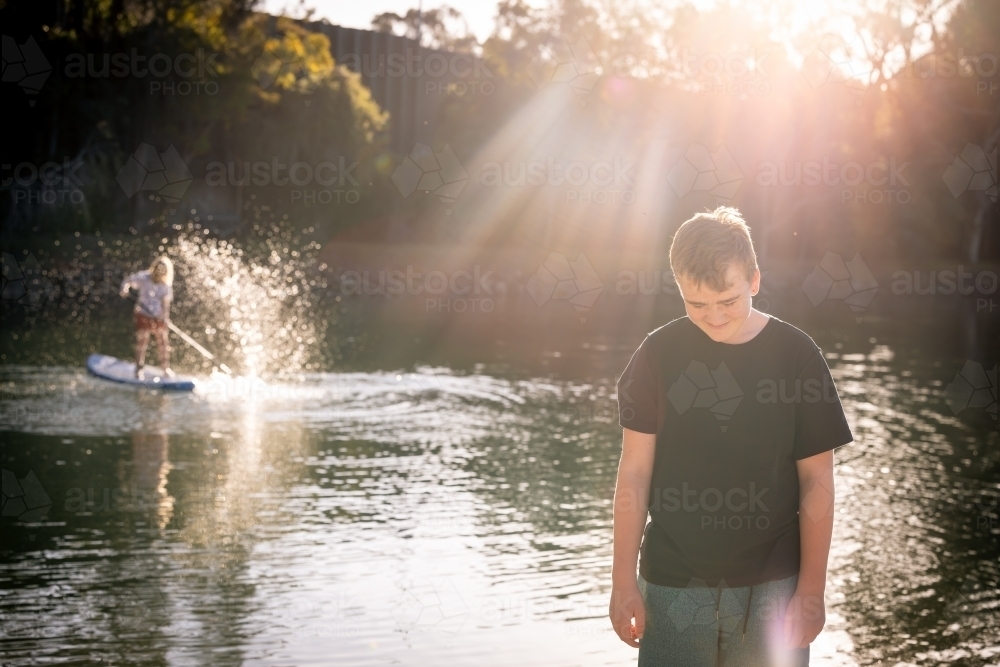 Teen boy in golden afternoon sunlight with brother splashing on stand up paddle board behind him - Australian Stock Image