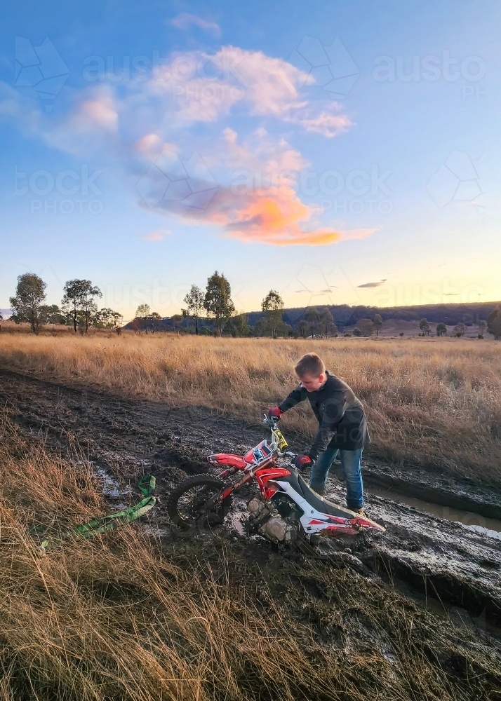 Teen boy on farm with motorbike stuck in the mud - Australian Stock Image