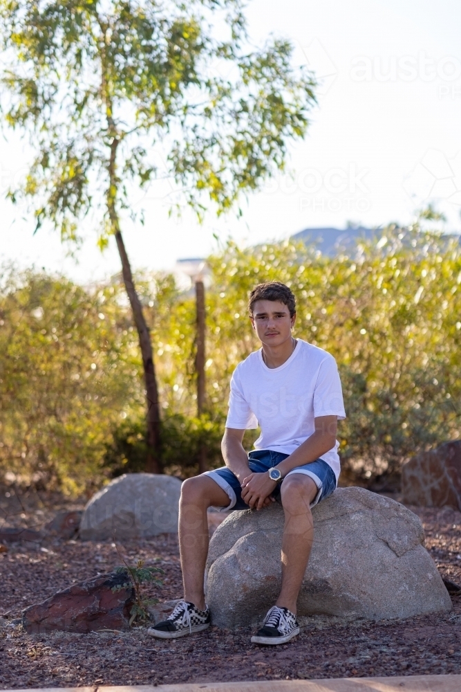 teen boy outdoors wearing white tee shirt sitting on rock - Australian Stock Image