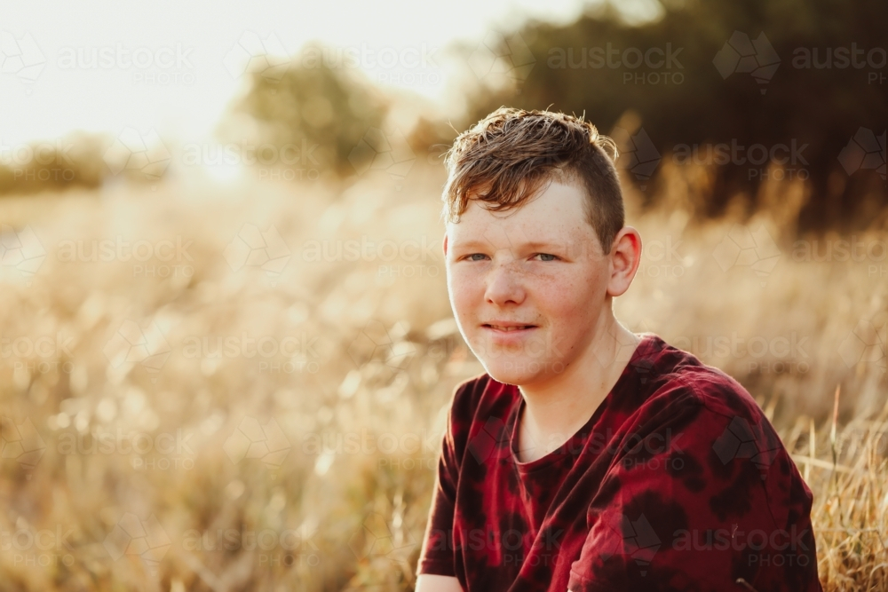 Teen boy sitting in long dry grass in golden afternoon light - Australian Stock Image