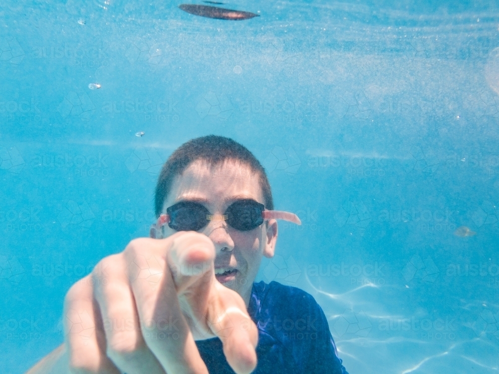 Teen boy swimming in pool underwater with goggles on pointing at you with hand - Australian Stock Image