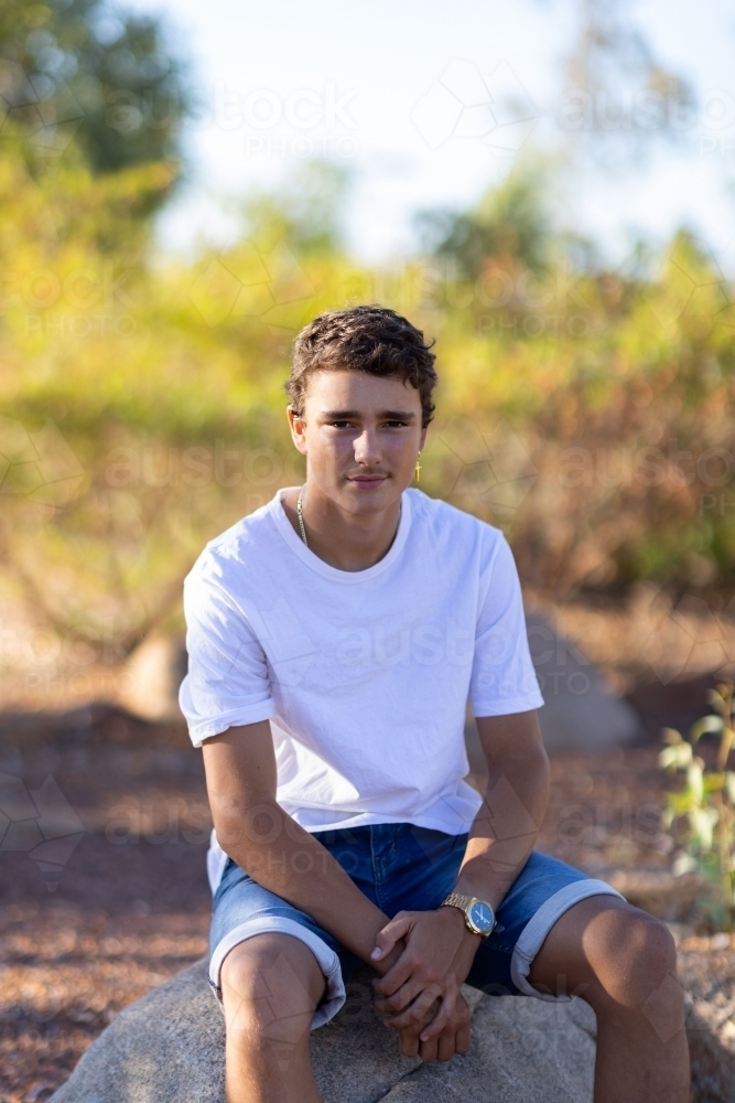teen boy wearing white tee shirt sitting on rock - Australian Stock Image