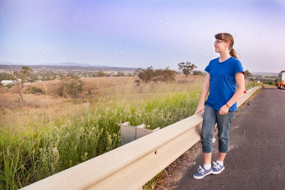 Teen girl by the roadside looking out over paddocks at dusk - Australian Stock Image
