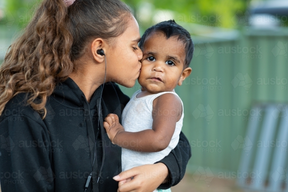 teen girl cuddling infant and kissing him on cheek - Australian Stock Image