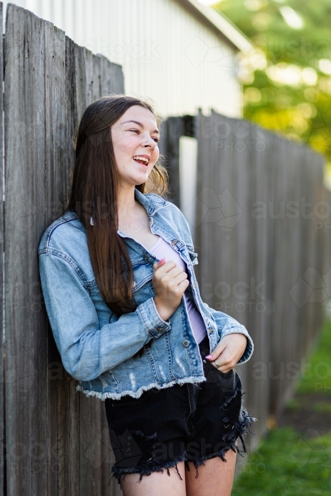 Teen girl laughing leaning on wooden fence in afternoon - Australian Stock Image