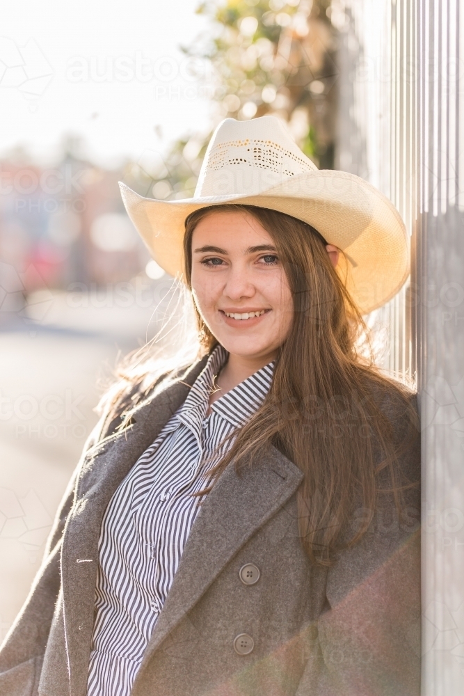 Teen girl leaning against fence smiling wearing hat - Australian Stock Image