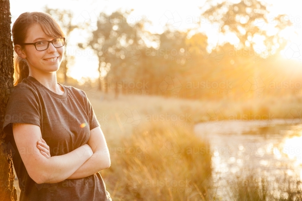 Teen girl leaning against tree with golden sun flare and copy space - Australian Stock Image