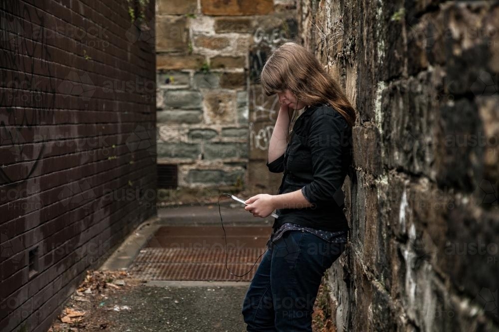 Teen girl leaning on alley wall listening to music - Australian Stock Image