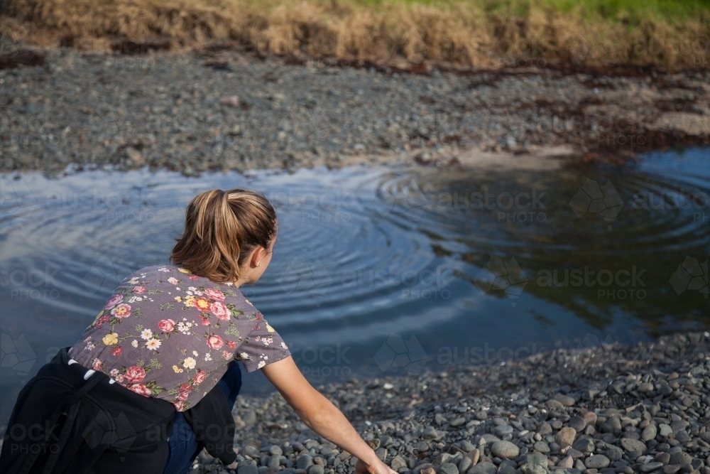 Teen girl picking up stones from beside water - Australian Stock Image
