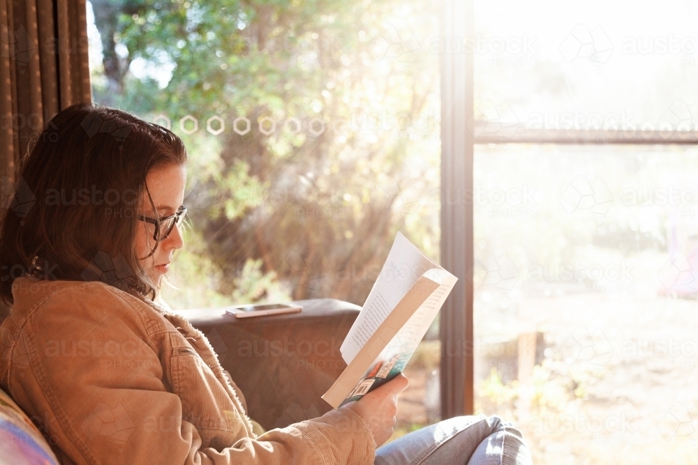 Teen girl reading a novel in the lounge room by a window in winter - Australian Stock Image