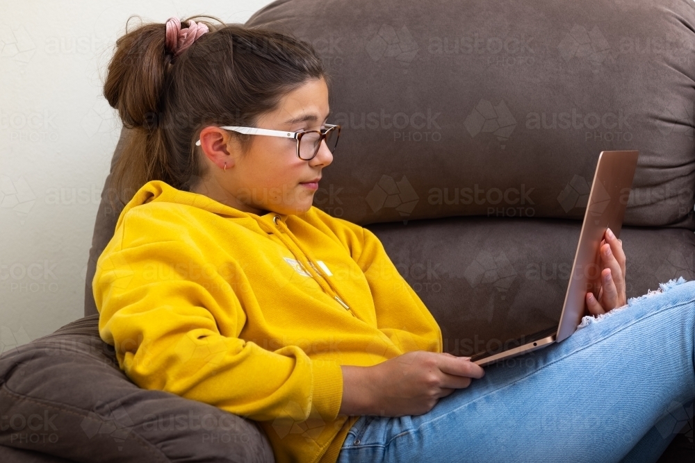 Teen girl wearing spectacles relaxing on sofa with laptop computer - Australian Stock Image