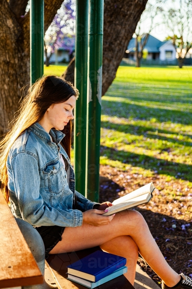Teen girl sitting at a park bench relaxing and reading a novel in the afternoon - Australian Stock Image