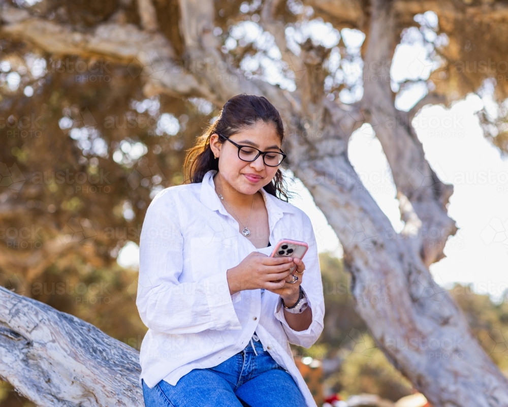 teen girl sitting in a tree with her smartphone - Australian Stock Image