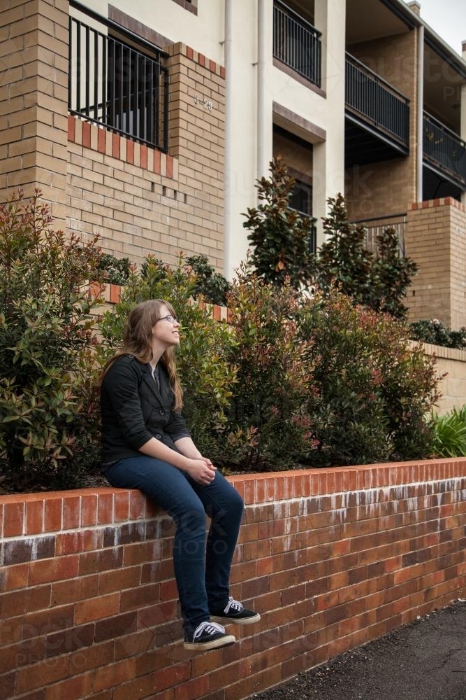 Teen girl sitting on brick wall of city house gardens - Australian Stock Image
