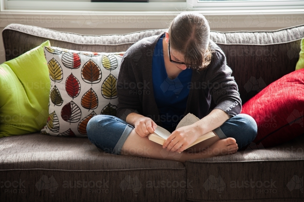 Teen girl sitting on lounge reading novel - Australian Stock Image