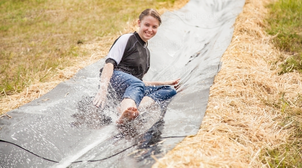 Teen girl sliding down a homemade waterslide laughing - Australian Stock Image