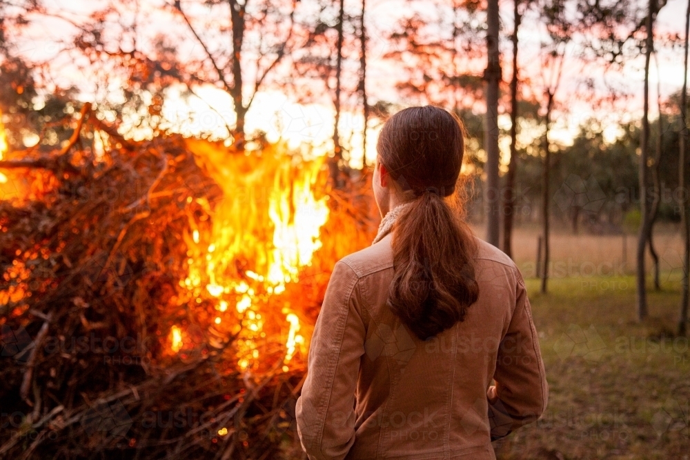 Teen girl standing by a bonfire in the back paddock at sunset - Australian Stock Image
