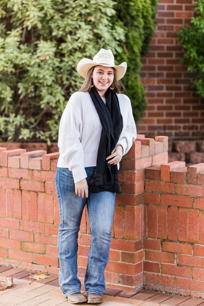 Teen girl standing leaning on brick wall smiling wearing hat and beanie - Australian Stock Image