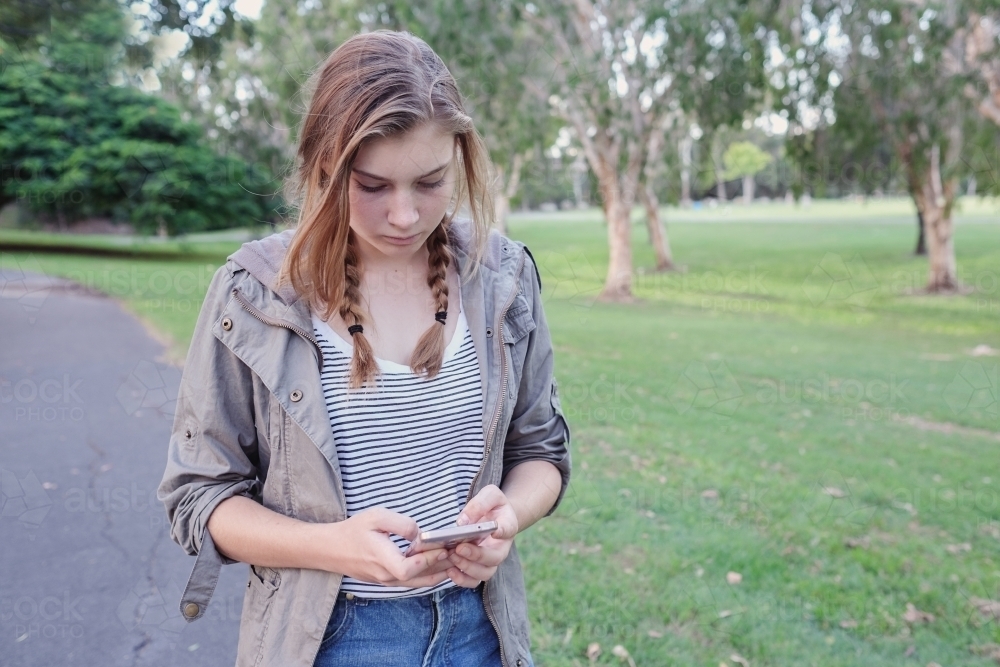 Teen girl using mobile phone while walking in the park - Australian Stock Image