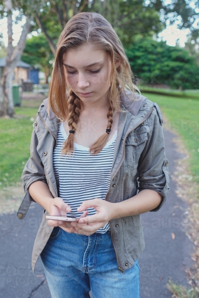 Teen girl using mobile phone while walking in the park - Australian Stock Image