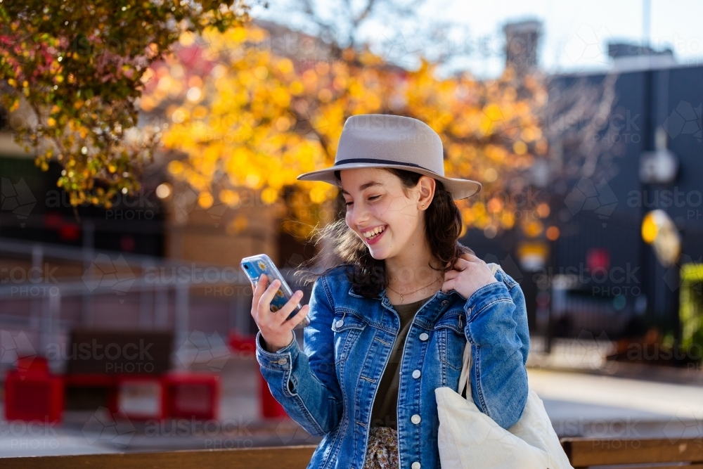Teen girl walking down street in country town looking at mobile phone on autumn afternoon - Australian Stock Image